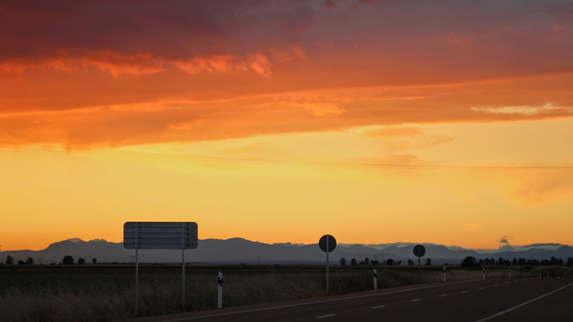 An empty road through flat fields under an orange sunset sky, with road signs silhouetted against distant mountains.