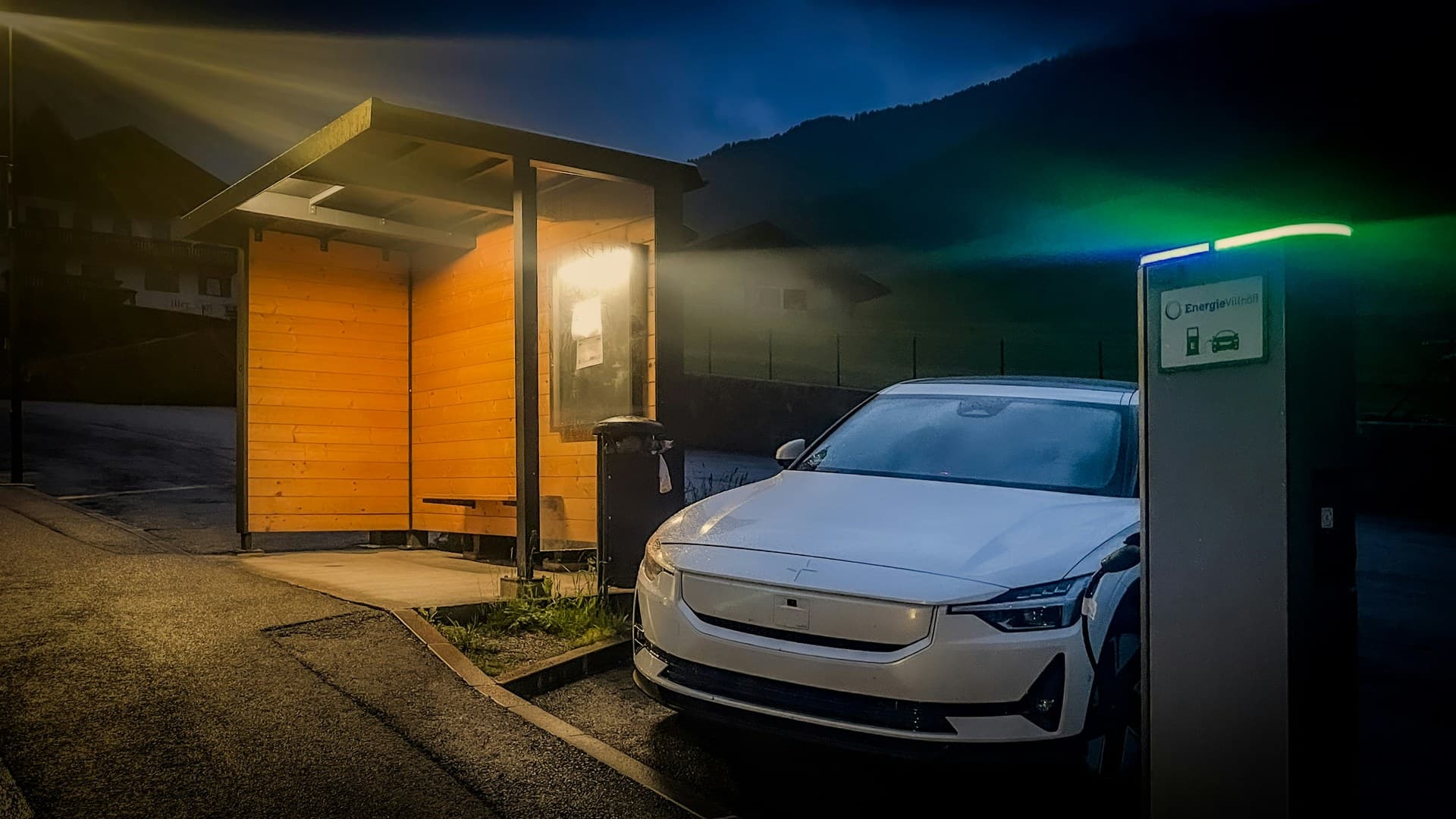 A white electric car plugged into a single charging post beside a wooden bus shelter in a mountain village at dusk.