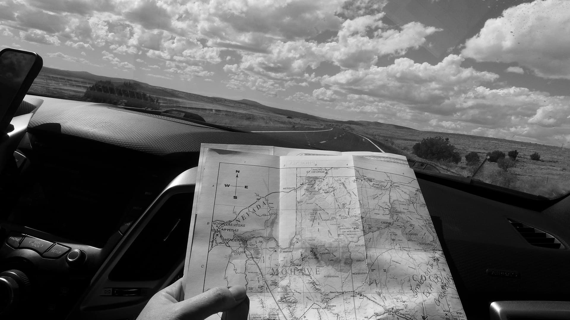 A black-and-white photo from a car's passenger seat: a hand holds a folded paper road map with handwritten notes over the dashboard, and an empty two-lane road runs across open desert under a cloudy sky.